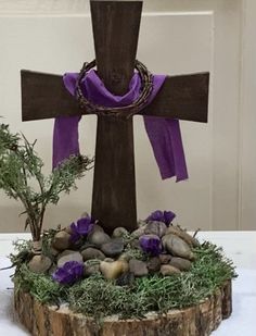 a wooden cross sitting on top of a table next to rocks and plants with purple ribbon