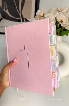 a hand holding a pink binder with the word t on it and some flowers in the background