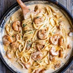 a skillet filled with pasta and shrimp on top of a wooden table next to a spoon