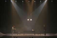 three people standing on stage in front of two spotlights and one person sitting at a piano