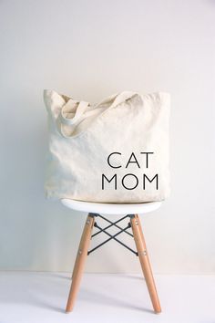 a cat mom tote bag sitting on top of a white chair with wooden legs