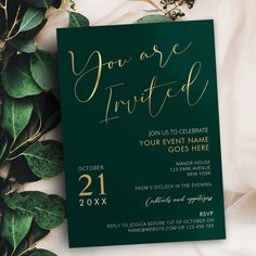 a green and gold wedding card with greenery around it on top of a white cloth