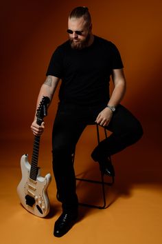 a man sitting on a chair with a guitar in his hand and wearing black pants