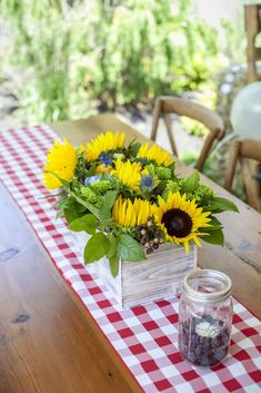 sunflowers and other flowers sit in a wooden box on a checkered tablecloth