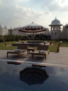 two lounge chairs and an umbrella on the edge of a swimming pool in front of a white building