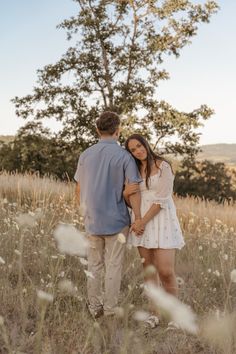 a man and woman standing in the middle of a field with tall grass, looking at each other