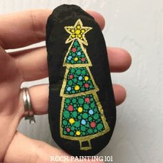 a hand holding a painted rock with a christmas tree on it and gold trimmings