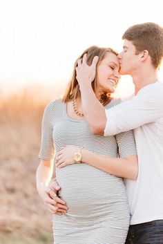 a pregnant couple cuddles and kisses while the sun is setting in the background