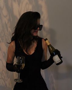 a woman in black dress holding a wine glass and bottle next to a white wall