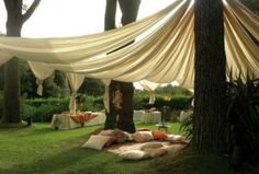 an outdoor wedding setup with draping and pillows on the grass, in front of trees