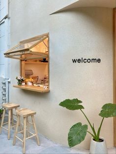 an open window on the side of a building with two stools and a potted plant