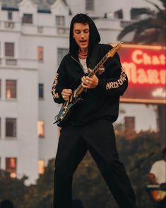 a man in a black hoodie is playing an electric guitar on stage with buildings behind him