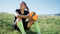 a woman sitting in the grass holding an orange and blue soccer ball with her legs crossed