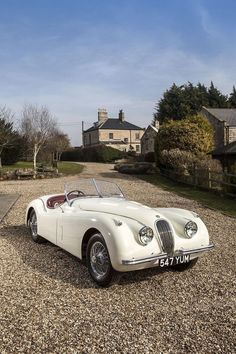 an old white sports car parked on gravel in front of a large house and driveway