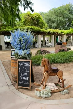 a wooden horse statue sitting next to a welcome sign and barrel with flowers in it