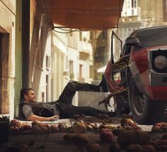 a man laying on the ground in front of a car with pumpkins around him