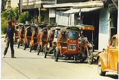 an old photo of people walking down the street in front of parked cars and tricycles