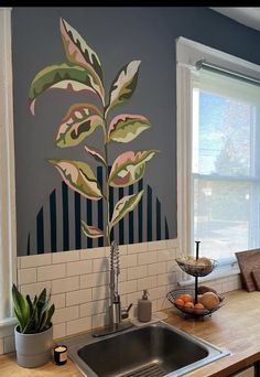 a kitchen sink under a window next to a painting on the wall and a potted plant