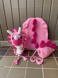 a pink crocheted dragon doll next to its bag and matching accessories on a tile floor