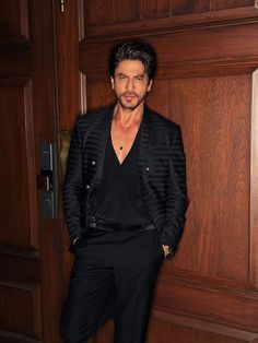 a man standing in front of a wooden door wearing a black suit and gold necklace