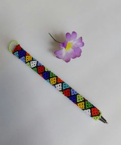 a colorful beaded bracelet next to a purple flower on a white surface with a needle