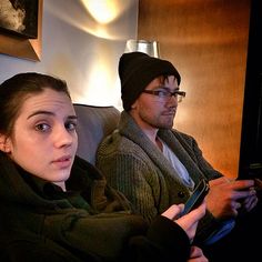 two people sitting on a couch with one holding a remote control and the other wearing a beanie