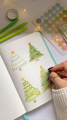 a person is drawing christmas trees on a book with markers and pencils next to it