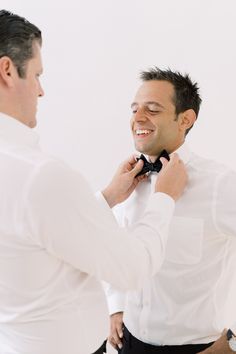 a man adjusting his bow tie while standing next to another man wearing a white shirt and black pants