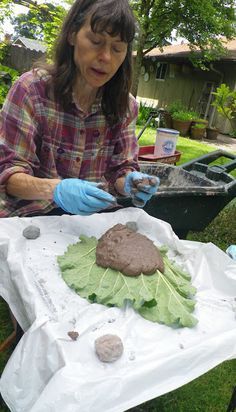 a woman in blue gloves is painting leaves and rocks on a sheet of white paper