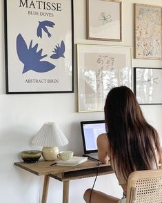 a woman sitting at a desk with a laptop computer in front of her and several framed pictures on the wall behind her