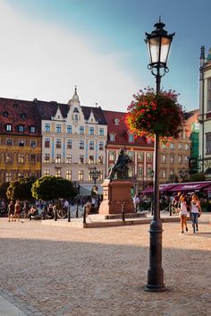 people are walking around in an old town square with many buildings and flowers on the lamp post