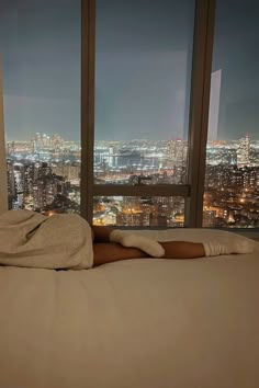 a woman laying on top of a bed in front of a window with city lights