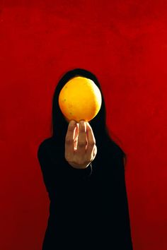 a woman holding an orange up to her face in front of a red wall,