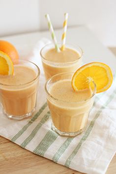 two glasses filled with orange smoothie on top of a wooden table next to an orange