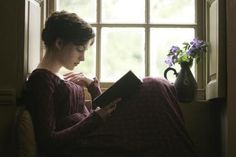 a woman sitting on a couch reading a book in front of a window with purple flowers