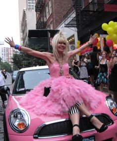 a woman in a pink dress sitting on top of a pink car with her arms outstretched