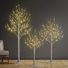 three lighted trees in the middle of a wooden floor next to a gray wall and chair