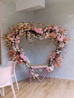 a heart shaped wreath with pink flowers and greenery on the wall next to a white chair