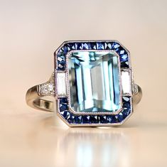 A stunning gemstone ring that centers on a 3.20 carat natural emerald-cut aquamarine. Baguette-cut diamonds and natural French-cut sapphires surround the aquamarine. Additional diamonds line the shoulders. This ring is platinum and displays an open-work under-gallery. ✦ GEMSTONE SPECIFICATIONS: Center Stone: Aquamarine Gemstone Weight: 3.20 Carats Gemstone Cut: Emerald Cut ✦ ENGAGEMENT RING SPECIFICATIONS: Stones: Aquamarine, Diamonds, Sapphires Material: Platinum Ring Style: Art Deco ✦ WHAT COM Diamond Ring With Halo, Emerald Cut Aquamarine Ring, Ring With Halo, Estate Diamond Jewelry, Emerald Cut Rings, Gemstone Engagement, Baguette Cut Diamond, Aquamarine Rings, Platinum Ring