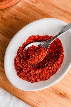 a white bowl filled with red spices on top of a wooden table next to a spoon