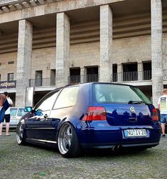 a blue vw golf wagon parked in front of a building with people standing around