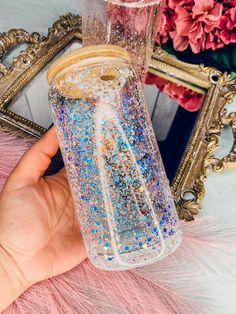 a hand holding a glass jar filled with blue and gold glitters next to a mirror