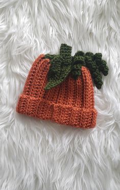 an orange knitted beanie with green leaves on top sitting on a white furry surface