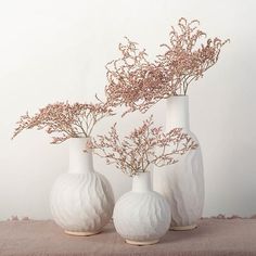 three white vases with pink flowers in them sitting on a cloth covered tablecloth