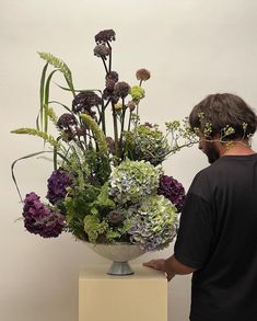 a man standing next to a large vase filled with purple, green and white flowers