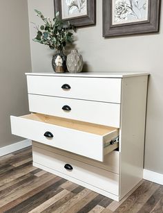 a white dresser with three drawers in front of two pictures on the wall above it