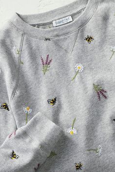 a grey sweatshirt with flowers and bees on the front is laying next to a white wall