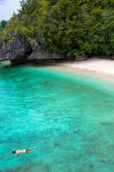 a person swimming in clear blue water next to a white sandy beach and green trees