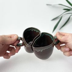 two hands holding small mugs in front of each other on a white surface with a plant behind them