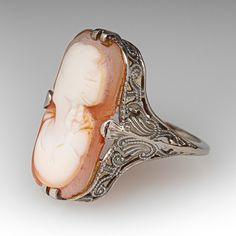 This antique filigree cameo ring features an elongated shell cameo of a woman's bust. The ring was loved and worn over the years and the cameo bust is a bit worn. The ring itself has crisp and strong details with open scroll work and milgrain. The ring is crafted in 10k white gold and is currently a size 5. The cameo is glued in for security. Vintage Filigree Intaglio Ring For Formal Occasions, Vintage Filigree Ring With Intaglio For Formal Occasions, Art Deco Wedding Jewelry With Cameo, Vintage Carved Filigree Ring For Formal Occasions, Antique Carved Filigree Ring For Formal Occasions, Art Deco Cameo Jewelry For Formal Occasions, Heirloom Filigree Ring With Cabochon For Wedding, Heirloom Filigree Wedding Ring With Cabochon, Oval Filigree Intaglio Ring For Wedding